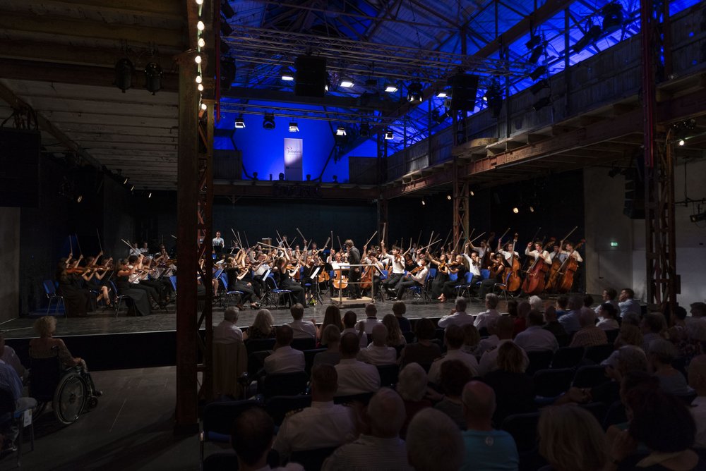 In einer alten Halle sitzen Personen auf Stühlen und schauen auf eine Bühne. Auf der Bühne ist ein Orchester mit klassischen Instrumenten.  
