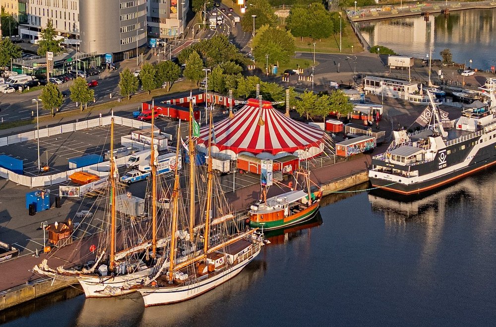 Luftaufnahme von Schiffen die an Kai kante liegen. Auf dem Größten Schiff ist die Aufschrift Koi und neben dem Schiff ist ein Zirkuszelt aufgebaut