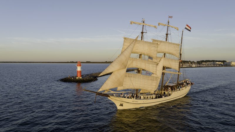 Das weiße Segelschiff Artemis fährt am roten Molenfeuer von Warnemünde auf die Ostsee zu einer Ausfahrt