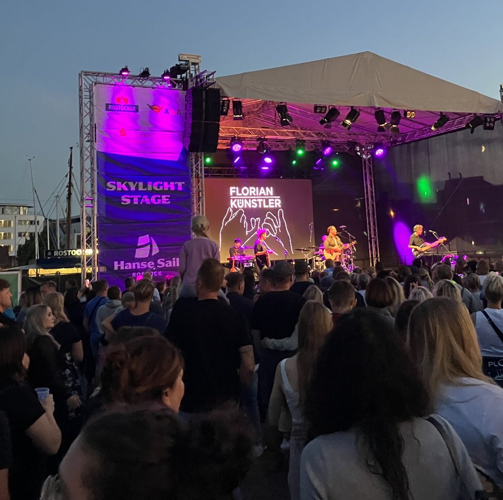 Vor Bühne stehen viele Personen. Auf Bühne steht eine Band. Auf Bannern steht Skylight Stage und Florian Künstler.