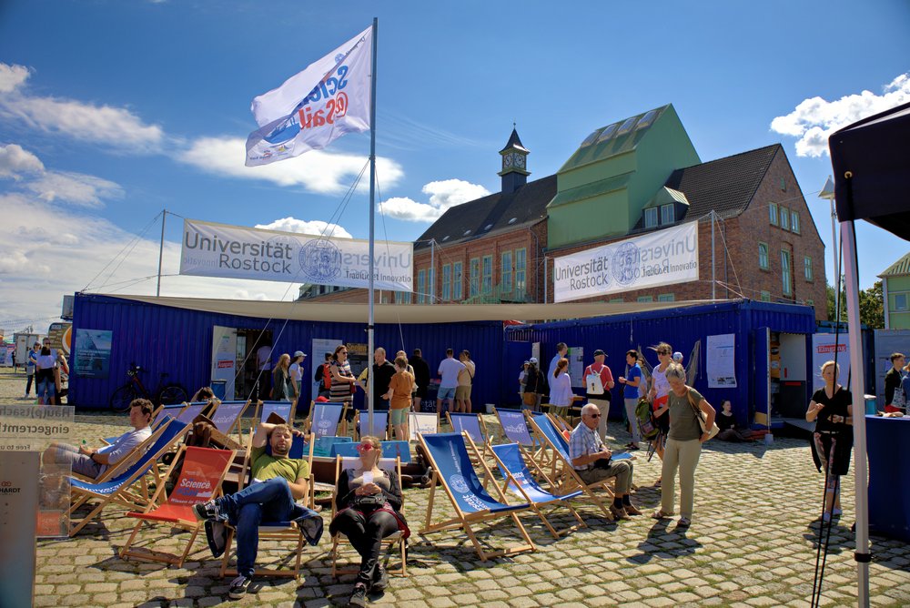 Männer und Frauen gemischten Alters, sitzen auf Strandstühlen vor Containern mit Universität Rostock Schriftzug