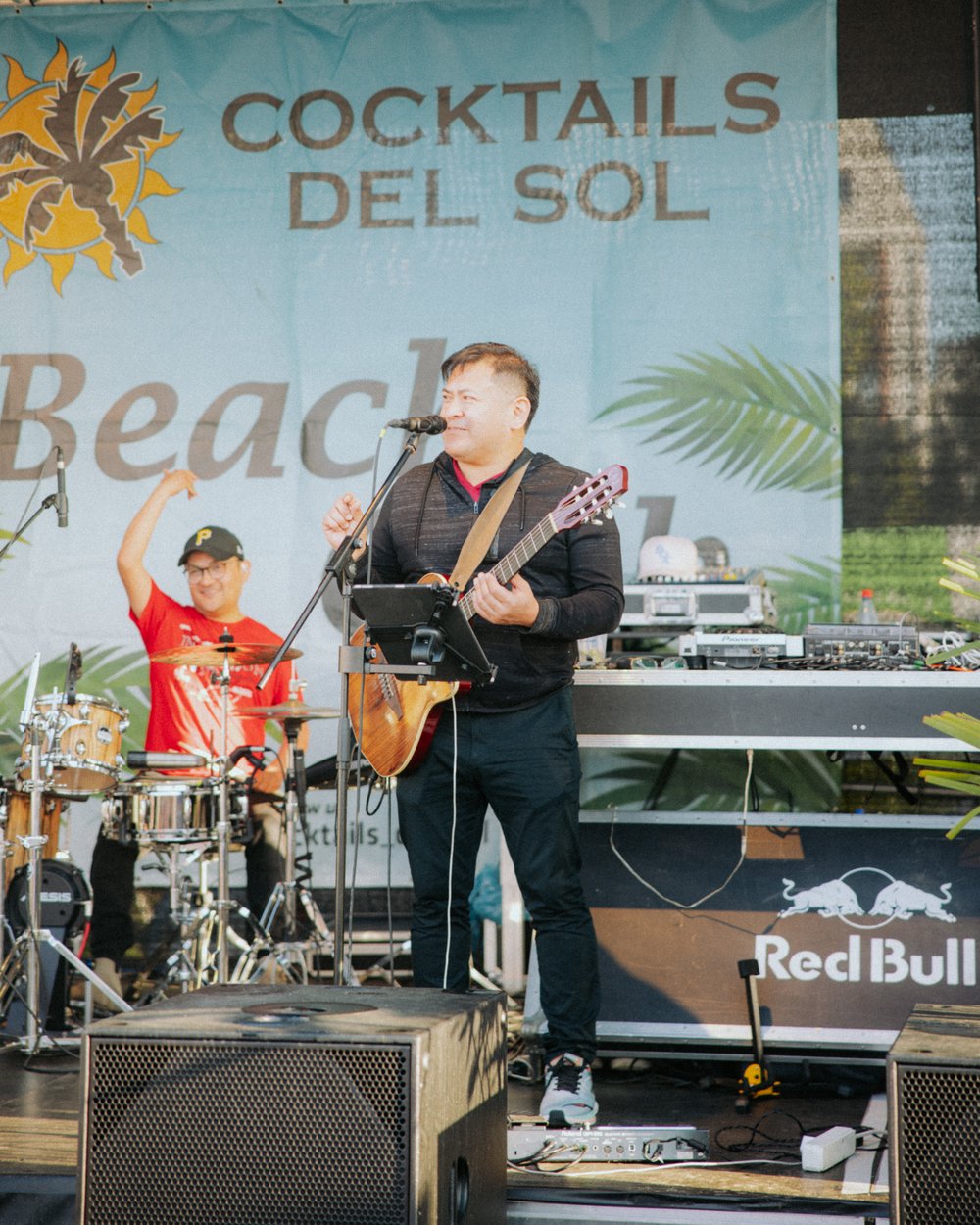 Dunkel gekleidete, männliche Person mit Gitarre steht auf Bühne. Hinter Ihm sitzt eine rot gekleidete, männliche Person hinter einem Schlagzeug. Auf einem Banner steht Cocktails del Sol.
