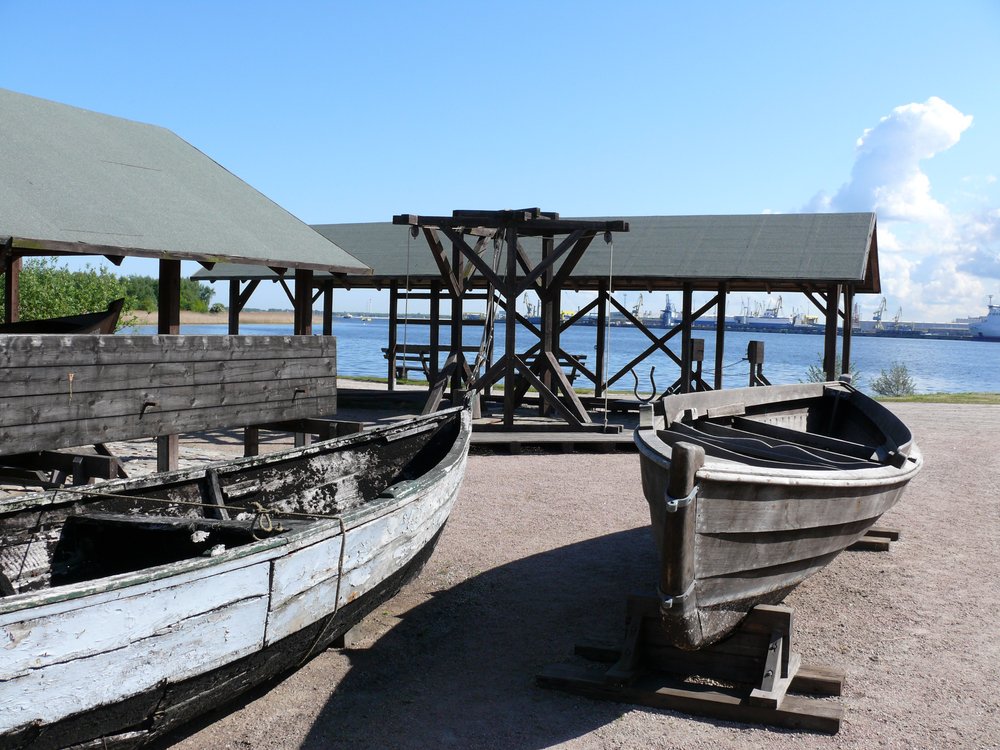 Alte Boote und Gebäude an Fluss im Vordergrund. Im Hintergrund auf anderer Flussseite moderne Hafenanlagen 