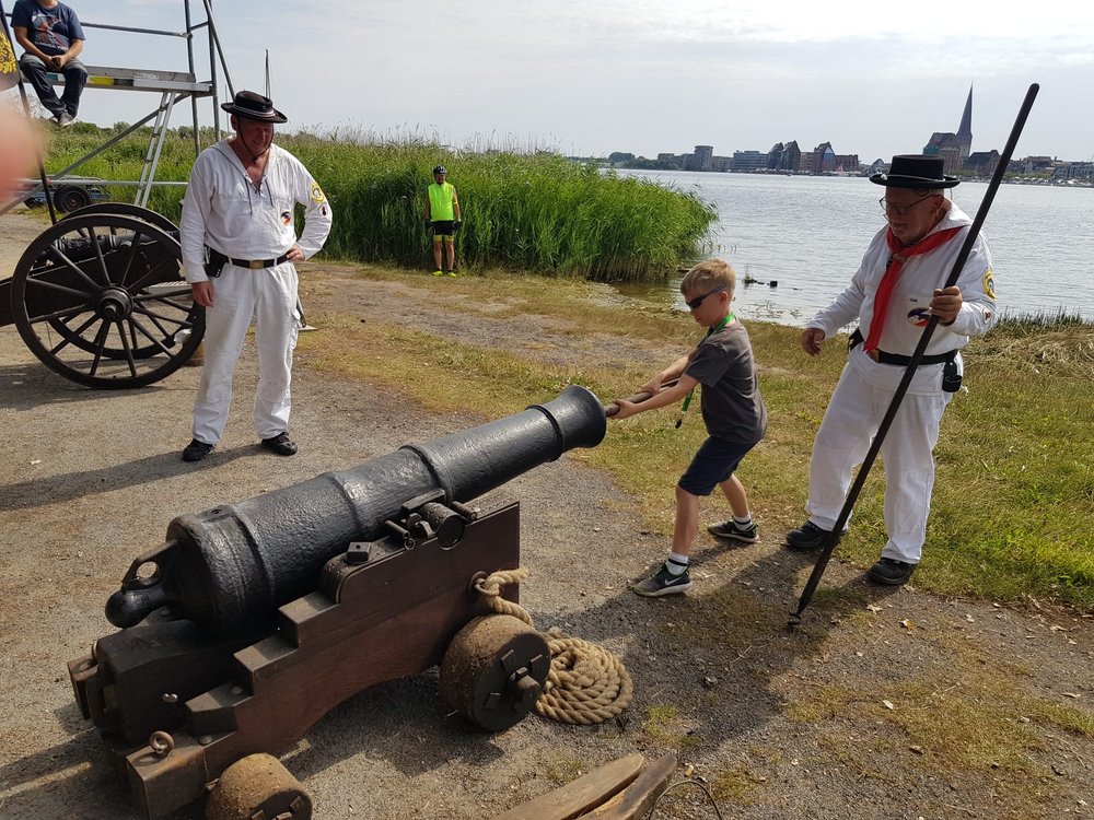 Kind ladet unter Aufsicht von Erwachsenen historische Kanone. Im Hintergrund der Fluss Warnow und die Rostocker Altstadt