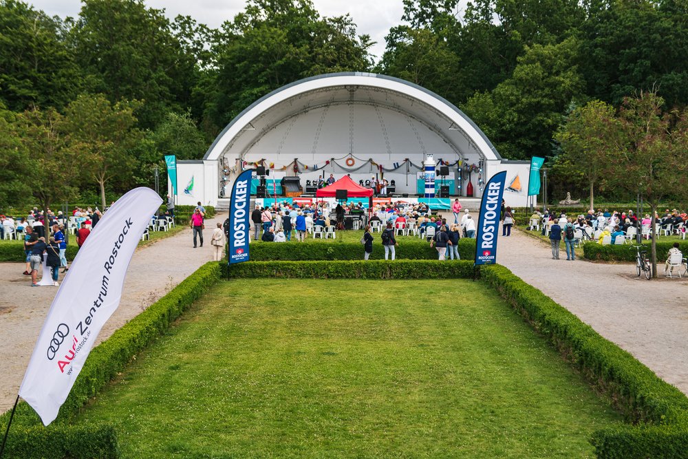 Blick auf die Bühne im Kurhausgarten Warnemünde. Vor der Bühne sitzen viele Personen auf Stühlen. Auf Werbefahnen steht Rostocker und Audi Zentrum Rostock