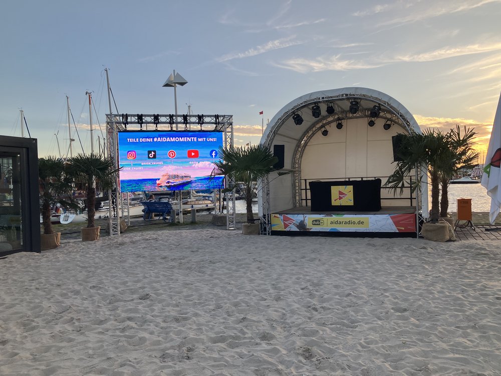 Auf Strandsand steht zwischen Palmen eine leere Bühne. Auf einer Leuchttafel neben  Bühne steht Aida. 